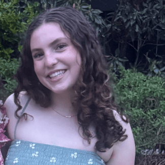 A smiling young woman with long, curly brown hair wears a sage green smocked top with a small floral print, standing in front of lush green foliage.