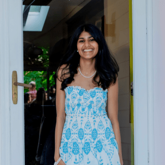 A smiling young woman with long dark hair, wearing a white strapless dress with a light blue geometric pattern and a pearl necklace, stands framing a doorway.