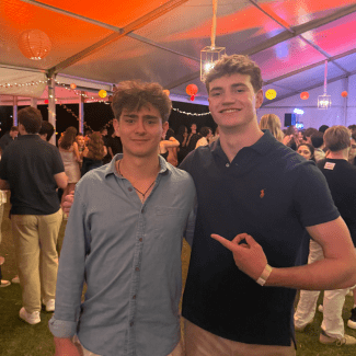 Two young men pose together under a lit event tent, with the person on the right pointing toward the person on the left who wears a light blue button-down shirt.