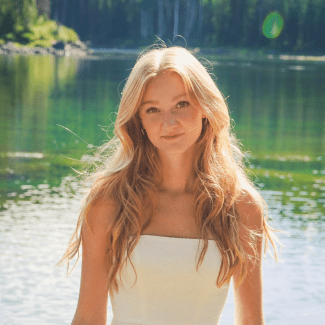 A young woman with long blonde hair, wearing a white strapless dress, poses in front of a calm, reflective body of water surrounded by trees.