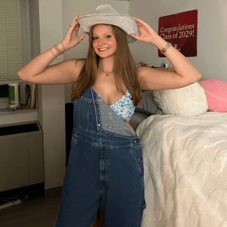 A smiling young woman poses in a room wearing denim overalls over a floral crop top, holding the brim of a sparkly silver cowboy hat.