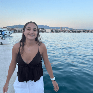 A smiling young woman with dark hair wears a dark sleeveless top and white pants while standing on a waterfront pier with boats and mountains in the background.