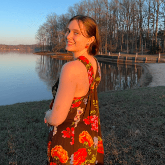 A young woman with short hair looks back smiling while wearing a floral-patterned dress in front of a quiet lake at sunset.