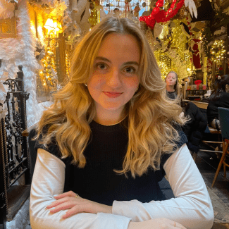 A smiling young woman with wavy blonde hair wears a dark sweater vest over a white long-sleeved shirt while seated at a table in a festively decorated restaurant.