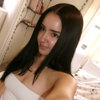 A young woman with long, straight black hair and a nose piercing smiles gently in a selfie while wearing a white strapless top.