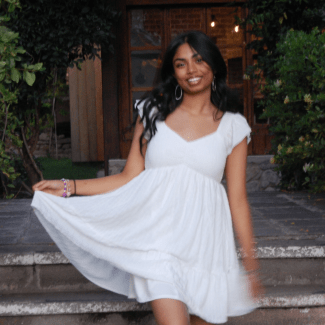 A smiling young woman with dark wavy hair twirls in a white tiered mini dress on stone steps outside a building.Nidhi Achanta