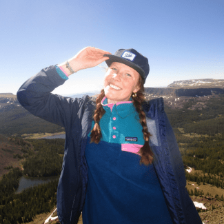 A smiling young woman with braided hair holds the brim of her baseball cap while standing on a mountain summit overlooking a valley.