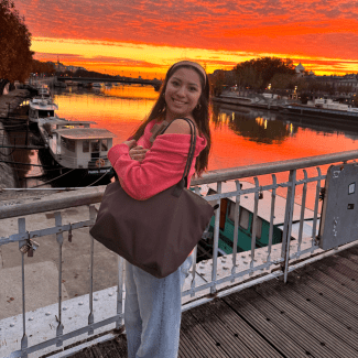 A smiling young woman wearing a pink sweater and holding a brown tote bag stands on a bridge overlooking a river under a bright orange sunset.