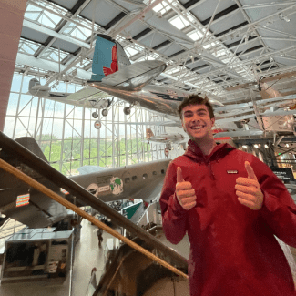 A smiling young man giving two thumbs up stands inside an aviation museum with vintage airplanes suspended overhead.