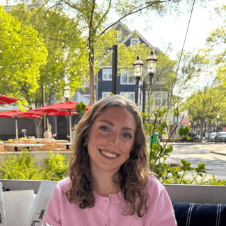 A smiling young woman with wavy hair wearing a pink sweater sits at an outdoor patio table in a sunlit town square.