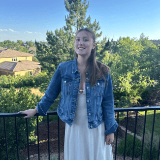 A smiling woman wearing a denim jacket over a beige dress stands on a balcony against a backdrop of trees and houses.