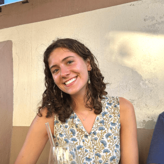A smiling young woman with dark curly hair sits at an outdoor table with a cocktail in front of a stucco wall.