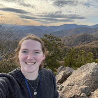 A smiling woman takes a outdoor selfie on a rocky mountain summit overlooking scenic valleys.
