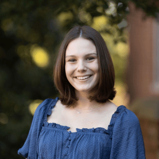 A outdoor portrait of a young woman with shoulder-length brown hair smiling broadly. She is wearing a blue ruffle-detailed blouse and standing against a softly blurred green foliage background.