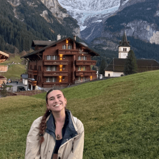 A smiling young woman with braided hair stands on a grassy hill in front of a large alpine chalet and mountains