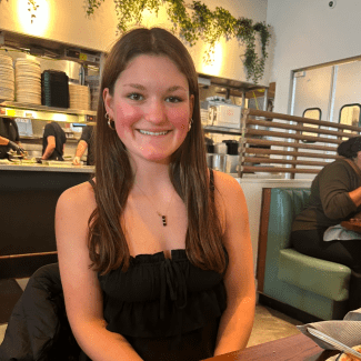 A smiling young woman with long brown hair, wearing gold earrings and a delicate pendant necklace, poses indoors with a restaurant kitchen area visible in the background.