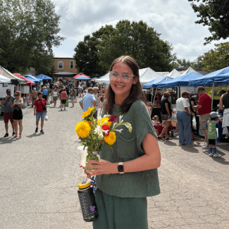 A smiling young woman with brown hair and clear-framed glasses holds a colorful bouquet of flowers at a bustling outdoor market with white and blue tents in the background.