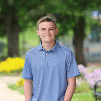 A smiling young man with short light brown hair, wearing a blue patterned polo shirt, poses outdoors with a soft-focus background of green trees.