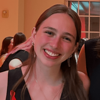 A smiling young woman with long brown hair, wearing a black top and earrings, tilts her head slightly toward the camera