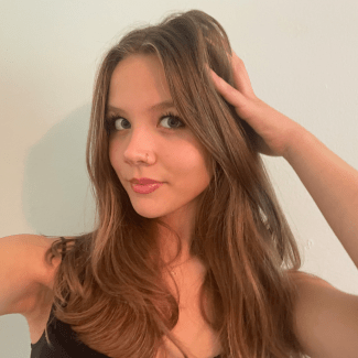 A young woman with long, layered brown hair posing for a selfie with one hand resting against her head in front of a neutral wall.