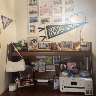A wooden dorm room desk with a hutch decorated with framed photos, art prints, stationery, a desk lamp, and a white printer.
