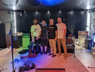 Four men stand arm-in-arm posing for a photo on a music stage equipped with amplifiers, microphones, and instruments.