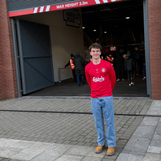 A young man in a red long-sleeved athletic jersey and blue jeans stands outside the entrance to a stadium.