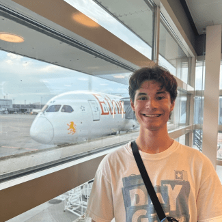 A smiling young man wearing a white graphic t-shirt stands near a window overlooking a large passenger airplane on the tarmac.