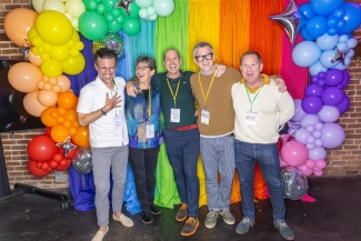 Five smiling people stand arm-in-arm posing for a photo in front of a rainbow-colored fabric backdrop flanked by vibrant balloon arches and mini disco balls.