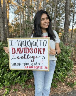 A smiling young woman holds a sign that reads: 'I matched to Davidson College! Thank you QB! Go Wildcats '28
