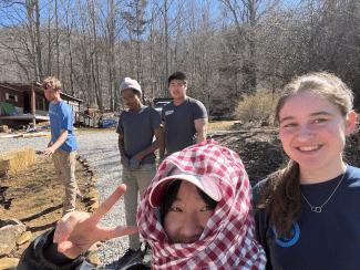 A group of young volunteers poses for an outdoor selfie on a sunny day, with a gravel path and wooded area in the background.