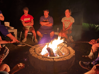 A group of people sits around a stone fire pit at night, roasting marshmallows with sticks over a bright, burning campfire.