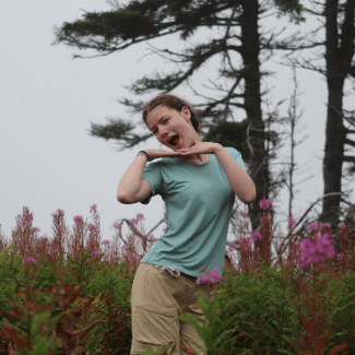 A young woman poses playfully with her hands under her chin in a field of pink wildflowers surrounded by evergreen trees.