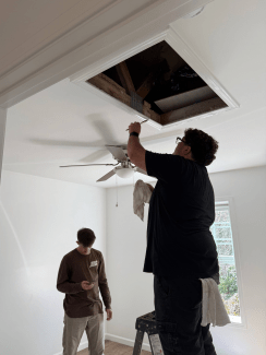 A volunteer standing on a step ladder paints white trim around an open ceiling attic hatch while another person looks down at a phone below.