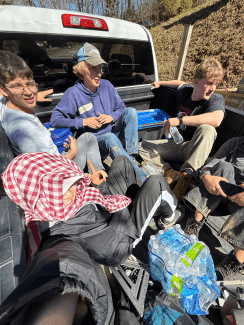 A group of young adults sits and chats together in the bed of a white pickup truck on a sunny day.