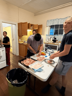 Several volunteers work together in a rustic kitchen to prepare a meal, chopping raw chicken and peppers at a white folding table.