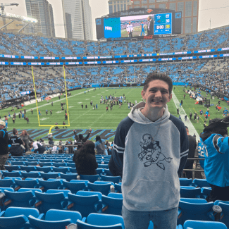 A smiling young man wearing a grey hoodie stands in the stands of a large outdoor football stadium filled with blue seats.