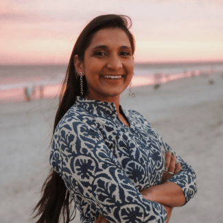 A smiling woman with long dark hair and crossed arms wearing a blue and white patterned tunic, standing on a sandy beach during a pink-hued sunset.