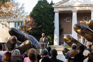 A speaker stands at a podium framed by large bronze hand sculptures, addressing a seated crowd outdoors.