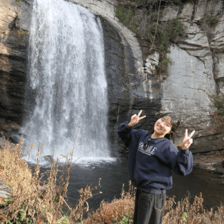 A smiling young woman in a dark sweatshirt flashes peace signs with both hands while posing in front of a tall waterfall cascading down a rocky cliff.