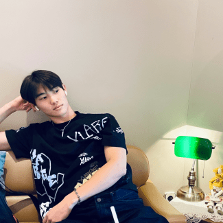 A young man in a black graphic t-shirt leans back on a leather couch beside a green banker's desk lamp.