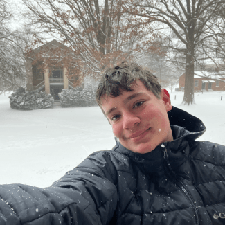 A smiling young man in a black hooded winter jacket takes a selfie outdoors in falling snow with a snow-covered campus building in the background.