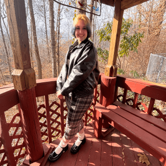 A smiling person with short two-toned blonde and blue hair stands inside a reddish-brown wooden gazebo wearing a black leather jacket and a plaid skirt.
