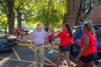 A man in a patterned short-sleeved button-down shirt and khakis high-fives a student wearing a red shirt and black shorts outdoors in a parking lot on move-in day. Other students in matching red shirts are visible in the background carrying boxes and supplies near a brick campus building.