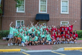 A large group of cheering college students posing together in front of a brick building. The group is divided into two distinct teams: one half wears matching light green t-shirts while waving and smiling, and the other half wears red Davidson Field Hockey shirts while posing with high energy.