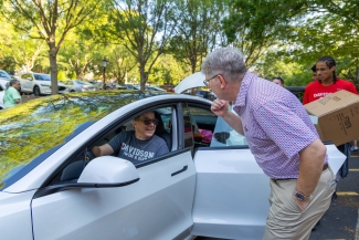 A man in a patterned short-sleeved shirt and khakis leans down to greet a driver sitting inside a white car during college move-in day. In the background, a student wearing a red Davidson shirt carries a large cardboard box alongside other parked vehicles under shady trees.