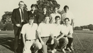 A black-and-white team photo of nine men posed outdoors on a golf course. Four members kneel in the front row, while four stand in the back row alongside an older man on the far left wearing a suit and tie.