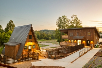 An A-frame cabin with warm exterior lighting sits beside a two-story wooden lodge connected by a paved, accessible ramp.