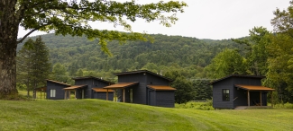Four dark wooden cabins with rusty-orange roofs sit lined up on a grassy hill before a dense green forest mountain.