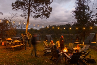 People gather around a blazing fire pit and picnic tables under string lights at dusk outdoors.